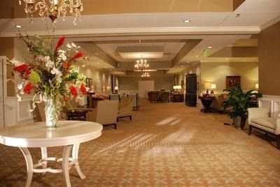 An elegant hotel lounge with a large floral arrangement on a white table. Light-colored carpet and furniture, with chandeliers.