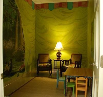 An interior room with green draped walls and a forest mural. Two armchairs sit by a lit lamp and small table with two chairs.