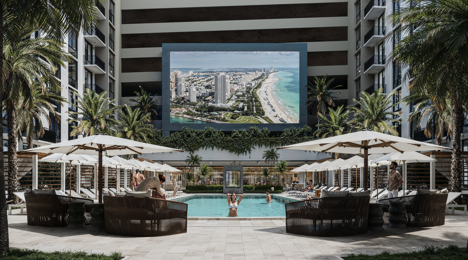 Swimming pool with lounge areas, large screen displaying beach view, palm trees, and hotel buildings.