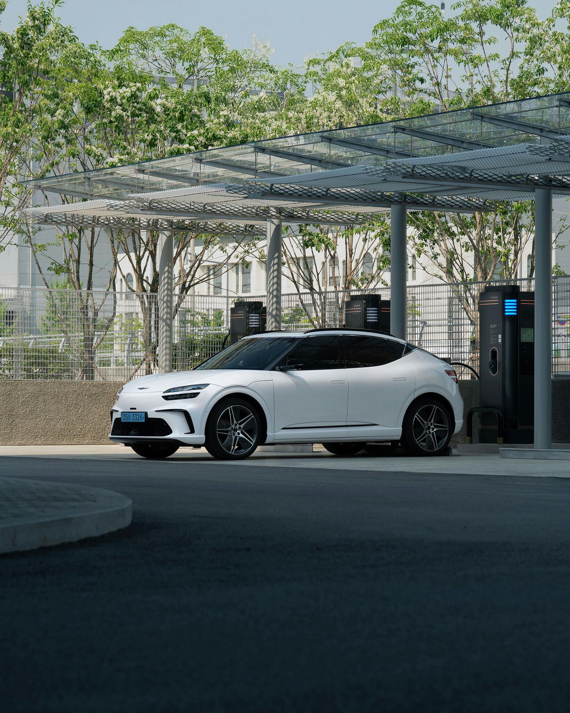 White electric SUV charging at a station under a canopy; trees in the background.