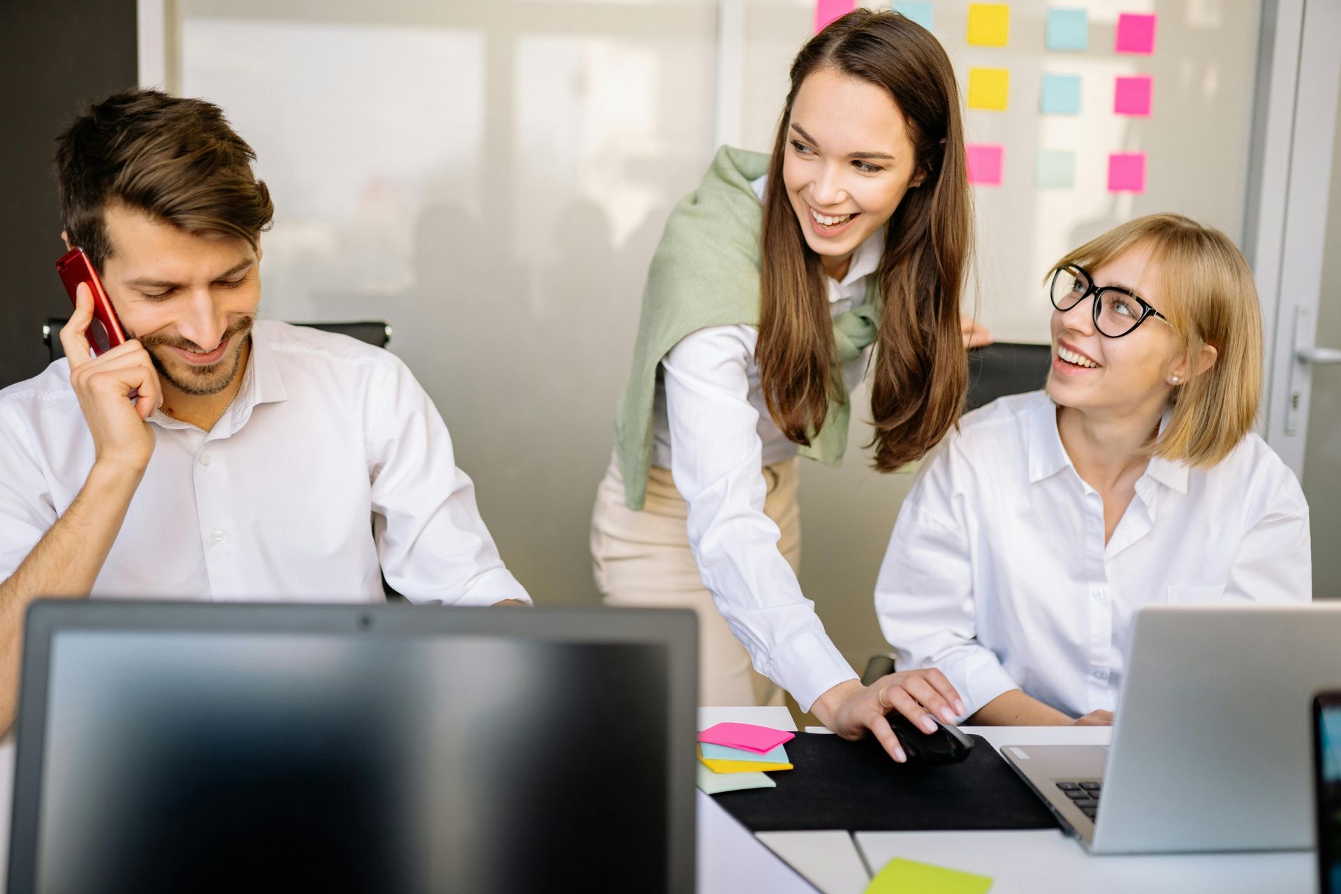Three coworkers at desks collaborating, one on phone, two looking at computer with smiles.
