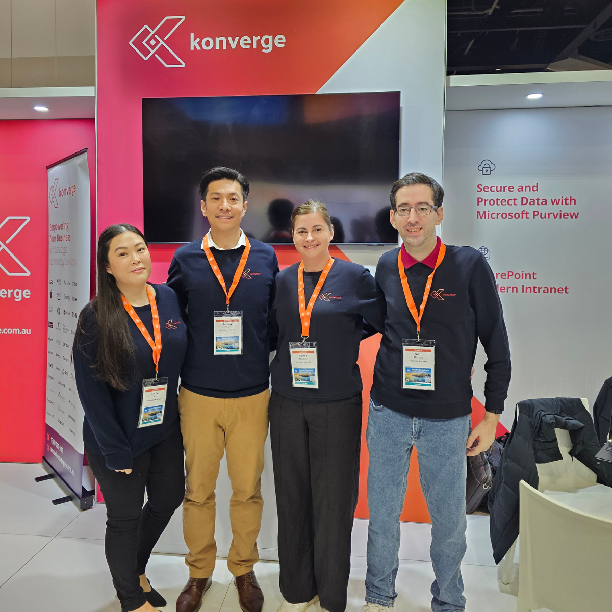Four people in matching blue shirts pose in front of a Konverge booth at a conference.