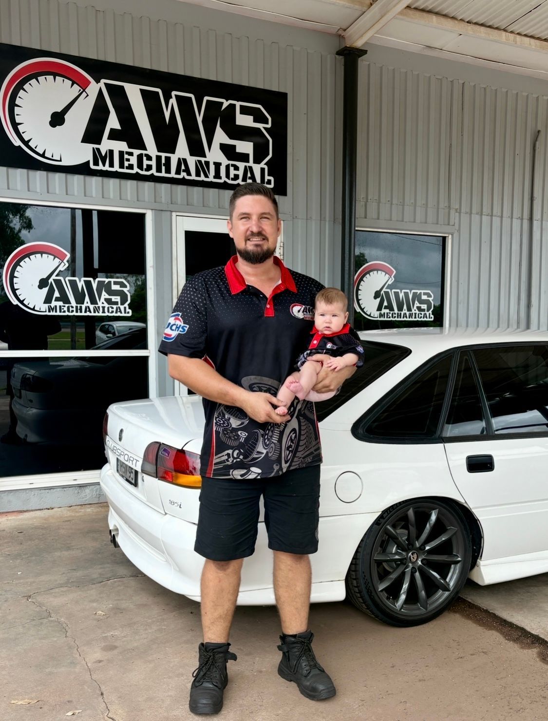 A man is standing in front of a white car holding a baby and smiling — AWS Mechanical in Stuart Park, NT