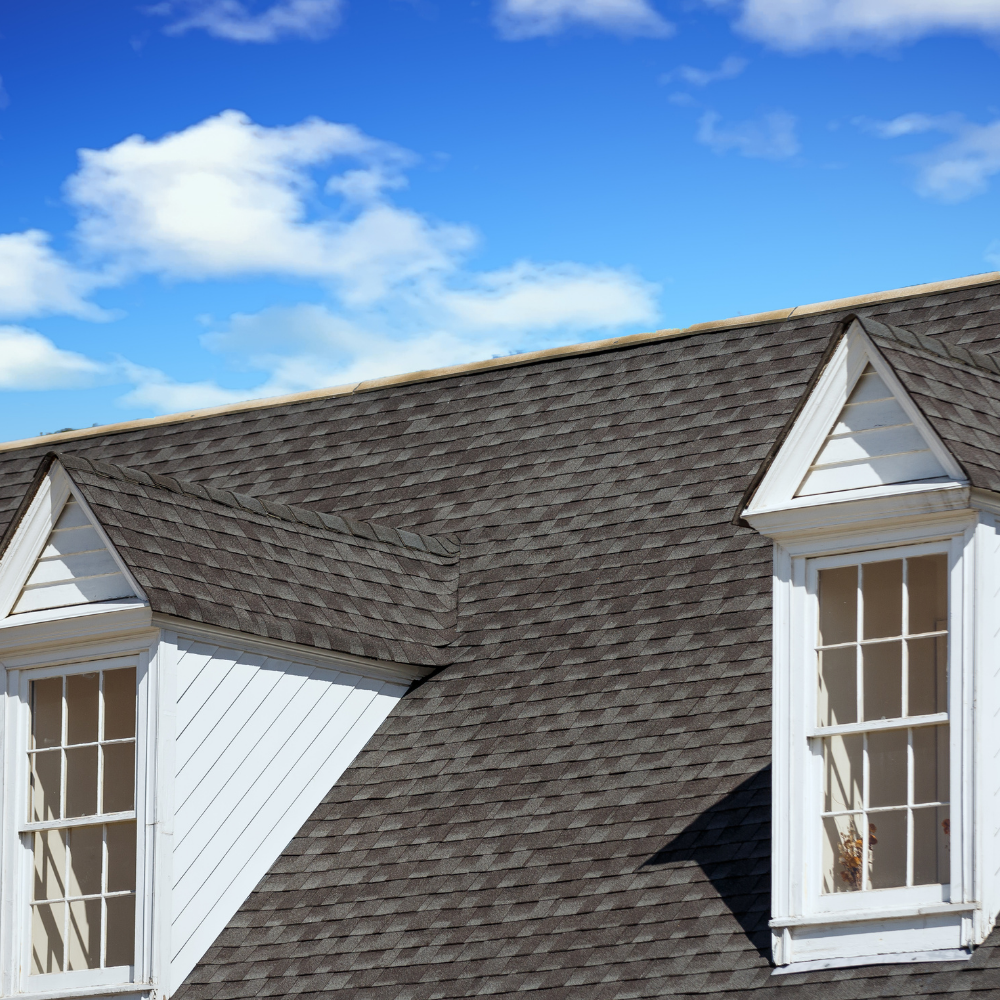 Gray shingled roof with two white dormer windows under a blue sky with clouds