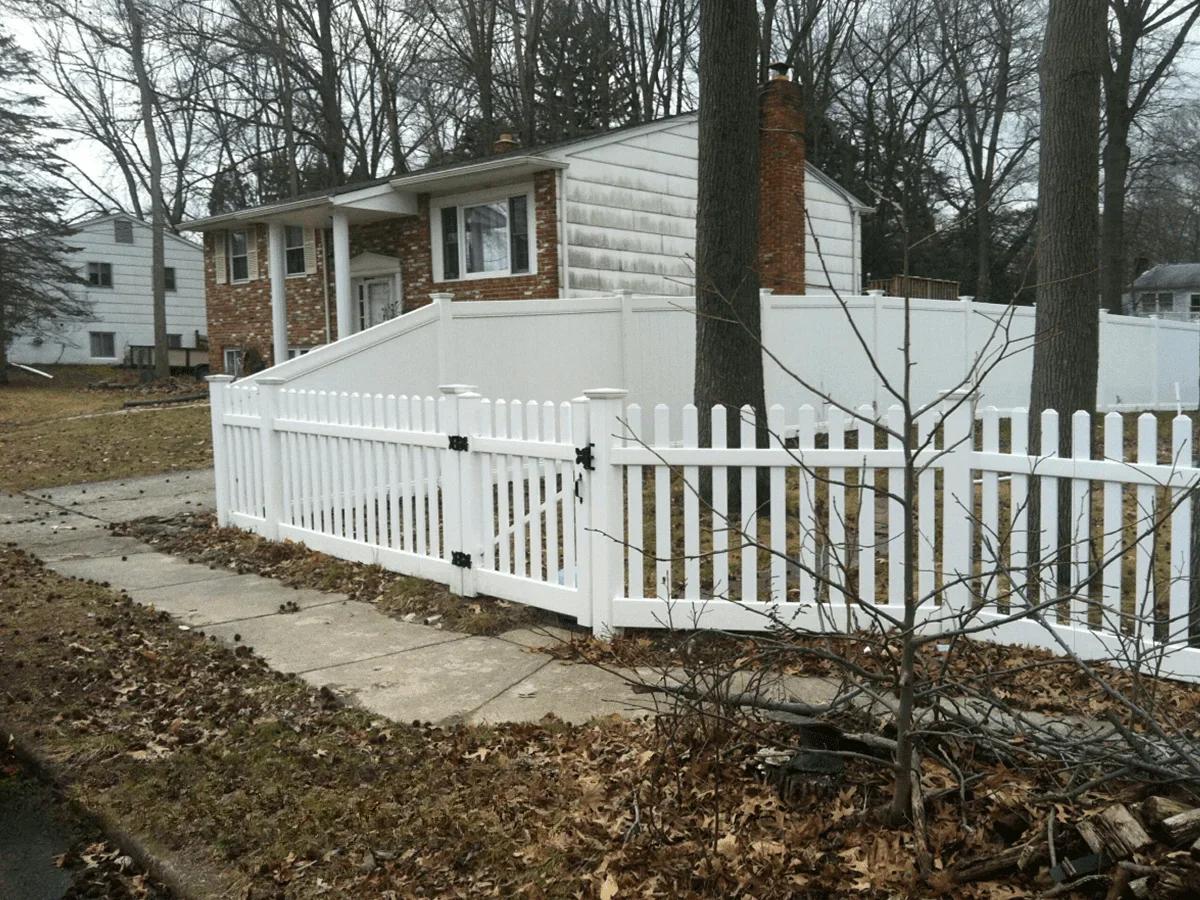 A white vinyl picket fence with a gate encloses a front yard with two trees in front of a house with stone siding.