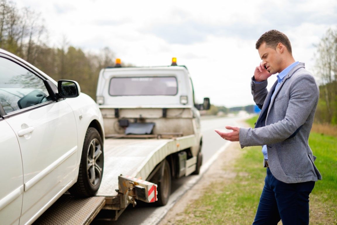 A person in a white shirt and jeans stands by their car with its hood up, talking on a phone in a grassy field.