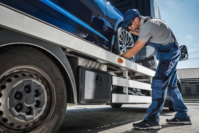 A worker in a blue uniform secures a vehicle wheel to a flatbed trailer using a metal chain.