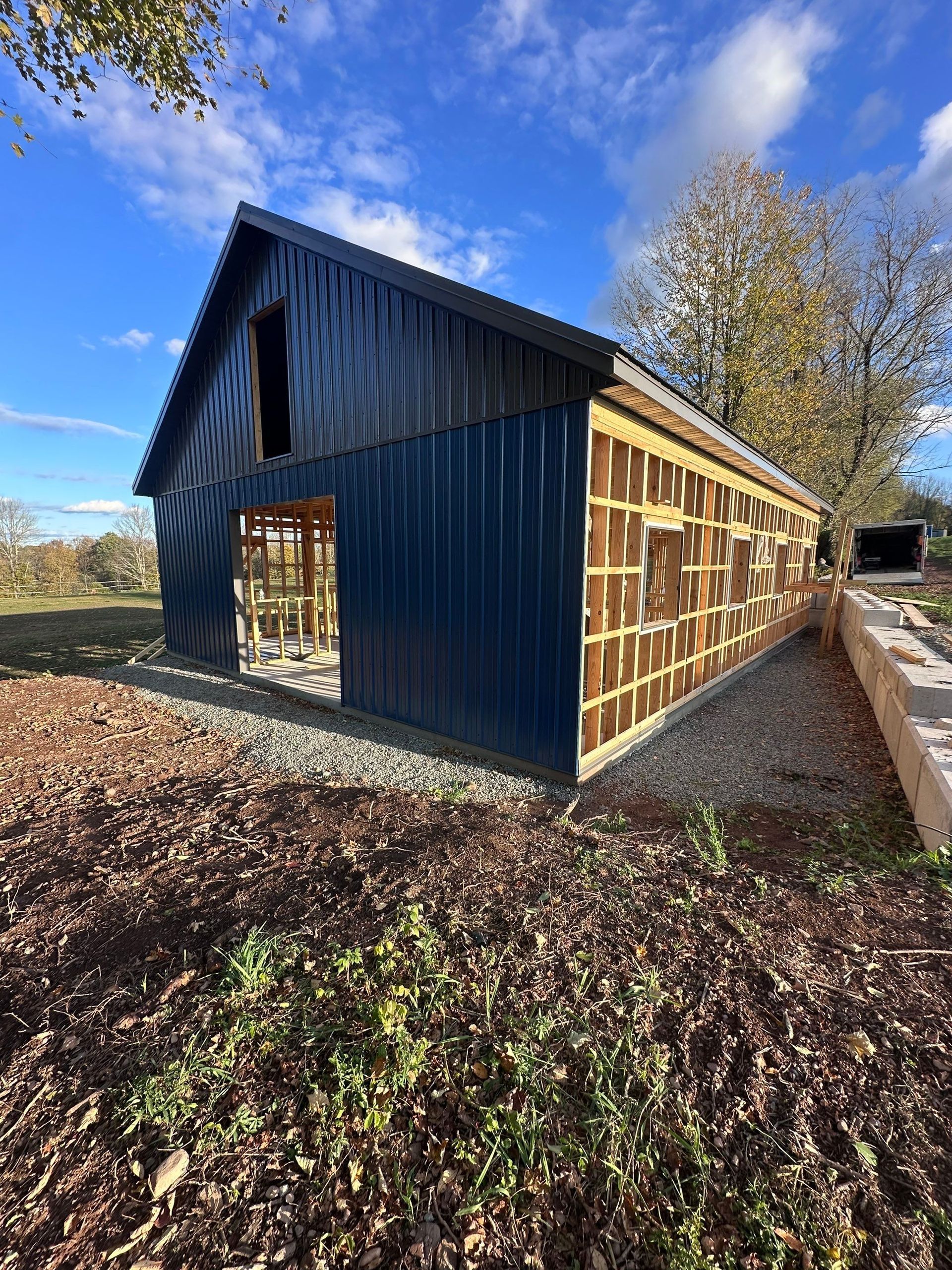 Dark blue wooden barn under construction on a grassy field beside a dirt road under a blue sky