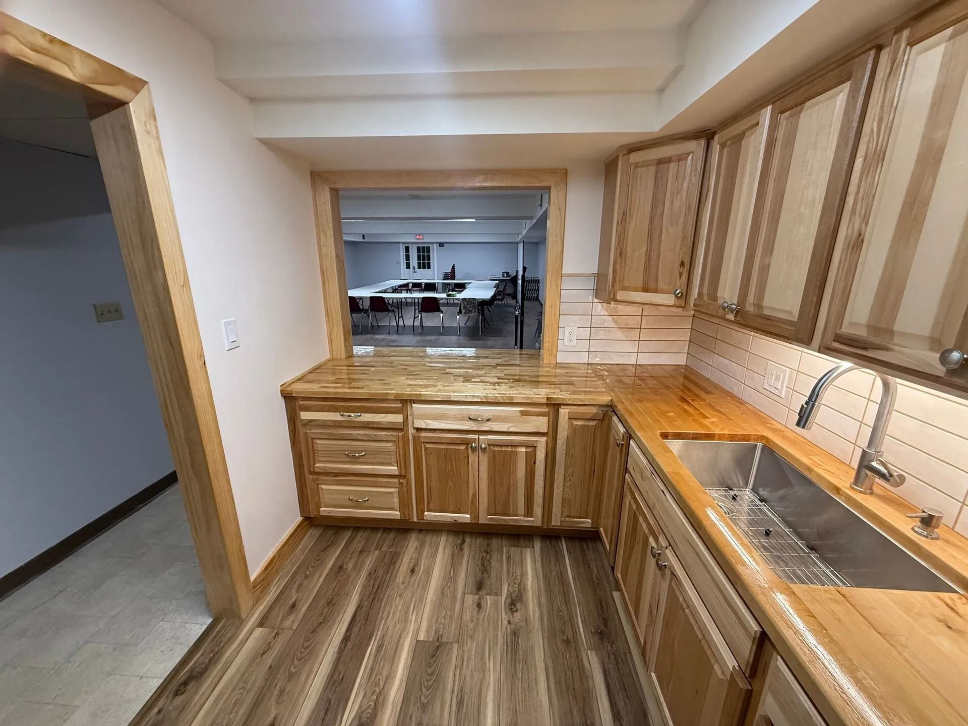 Wooden galley kitchen with cabinets, sink, and striped wood-look flooring, opening to another room.