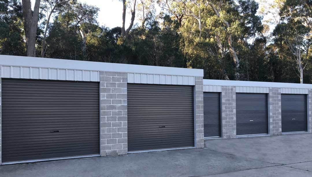 Storage units with gray metal roll-up doors, concrete block columns, and a metal roof, backed by trees. — Bay & Basin Storage In Huskisson, NSW