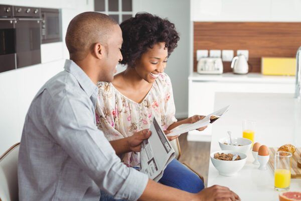 A couple sits at a kitchen table, smiling while looking at a newspaper and documents during breakfast.