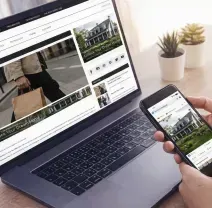 Person holding a smartphone while viewing a real estate website on a laptop in a desk workspace.