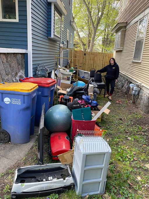 A woman is standing in front of a house surrounded by trash cans and boxes.