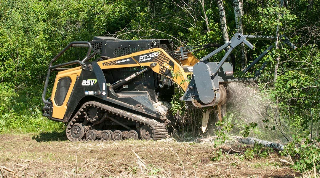 Yellow and black track loader with brush cutter clearing brush in a wooded area.