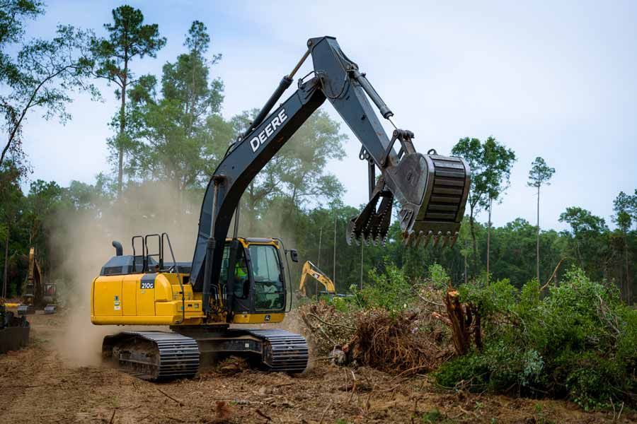 Yellow John Deere excavator clearing brush in a wooded area.