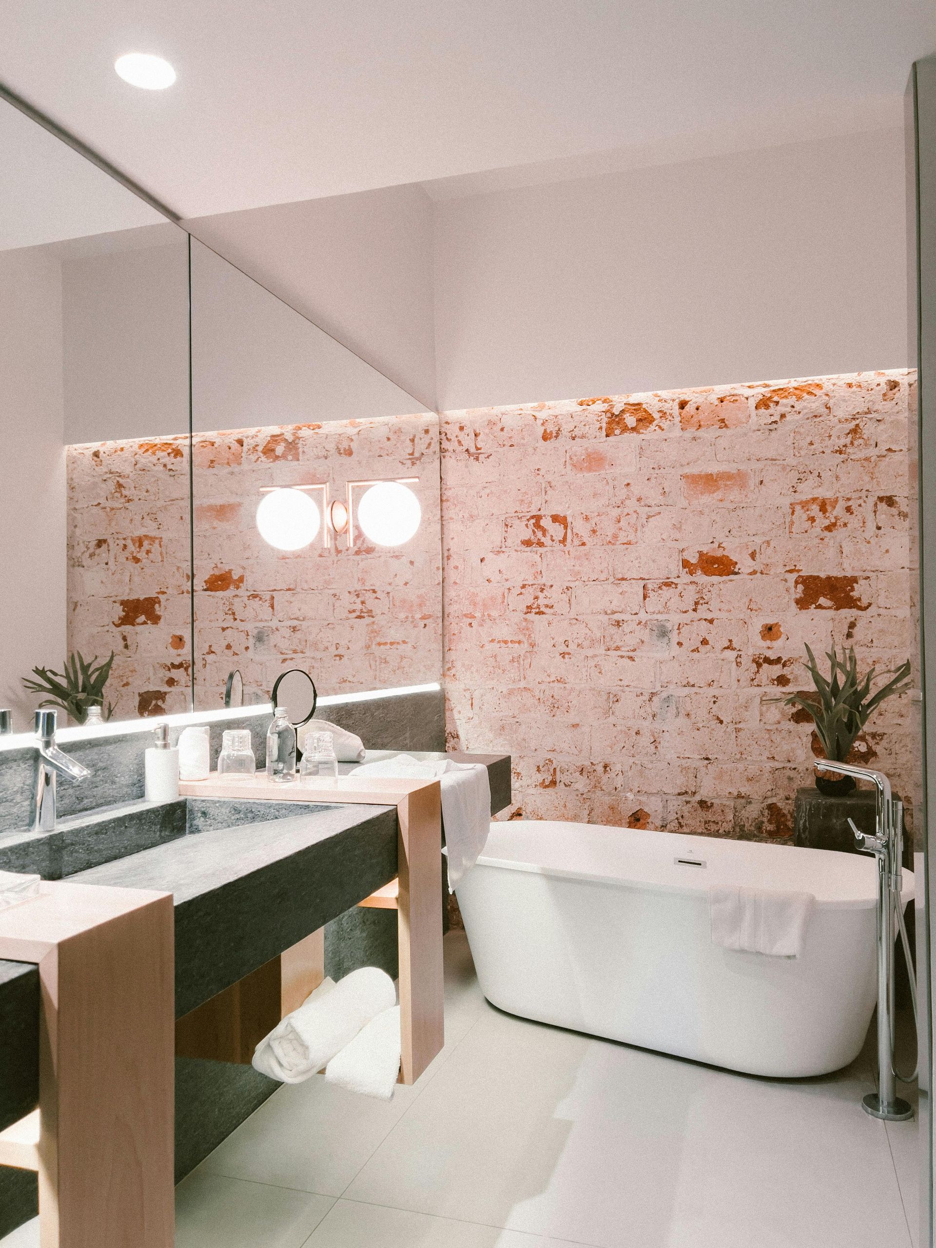 Modern bathroom with exposed brick wall, freestanding tub, double sinks, and large mirror.