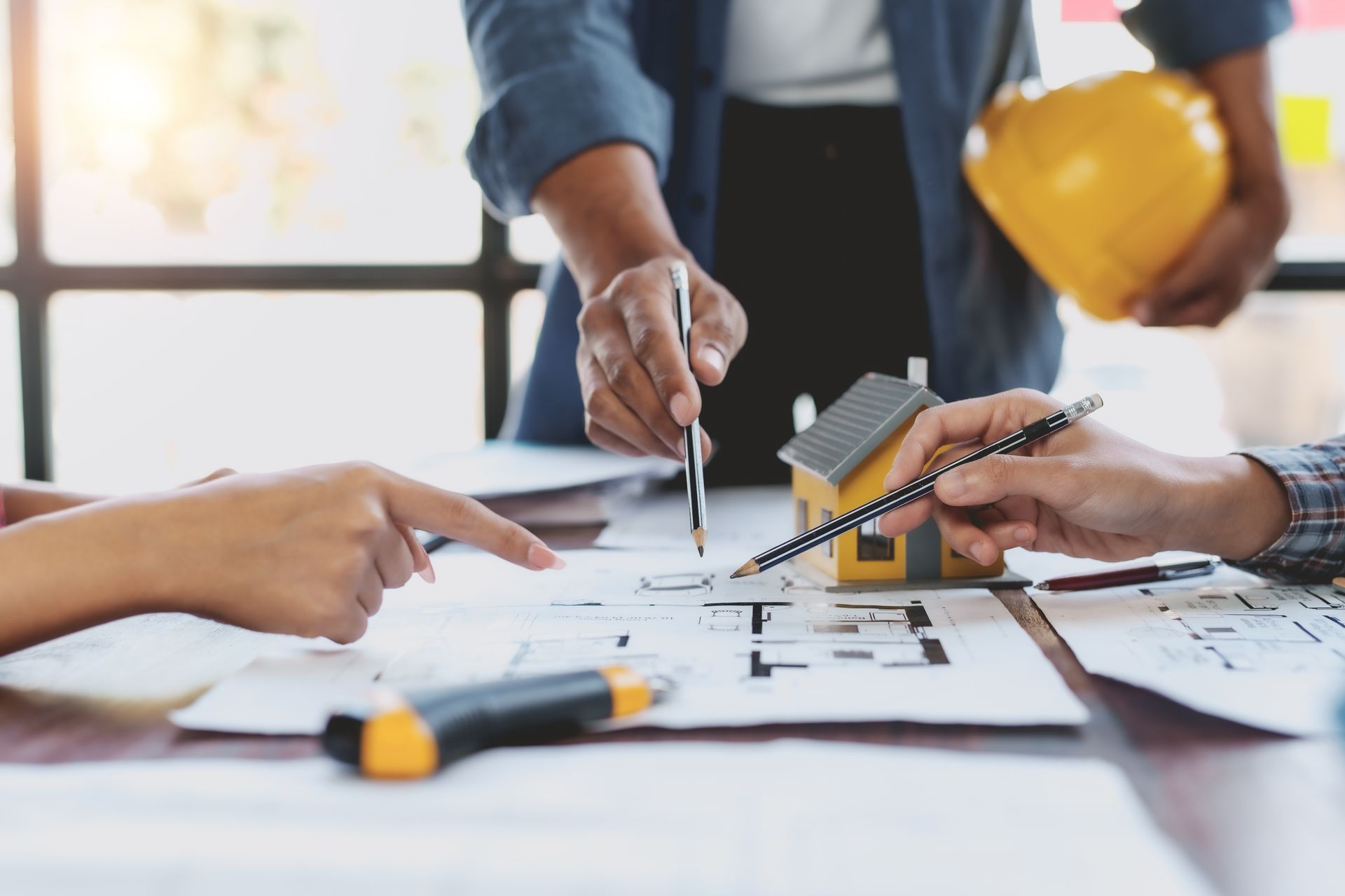 People collaborating on architectural plans at a desk, with a house model and hard hat.