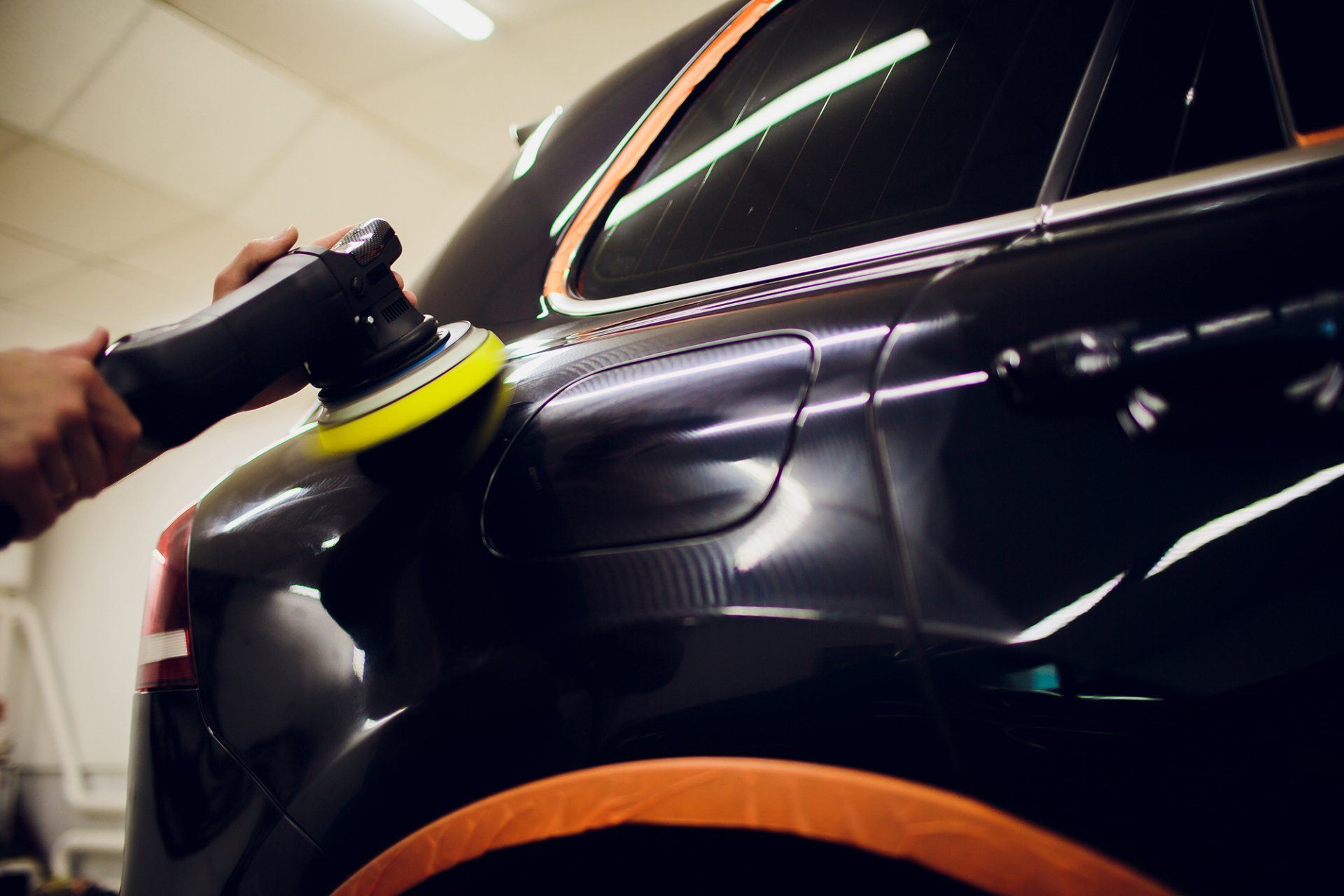 Persona che usa una lucidatrice elettrica sul pannello della carrozzeria di un'auto nera. Nastro arancione per proteggere le finiture.