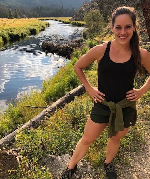 A woman in a black tank top and shorts is standing in front of a river.