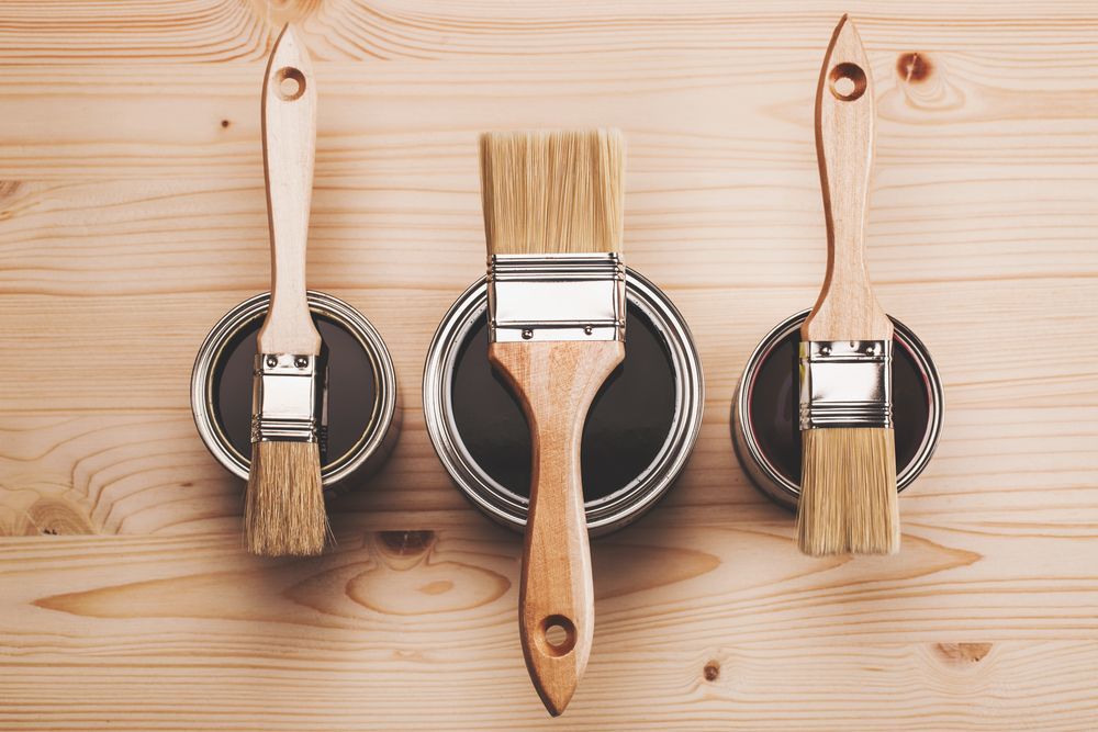 Three paint brushes are sitting next to cans of paint on a wooden table.