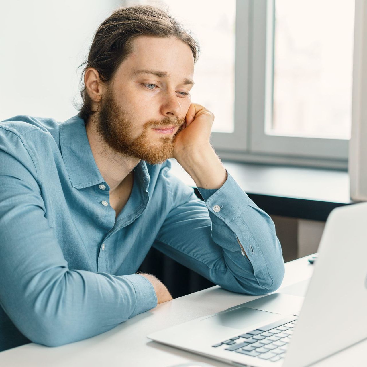 A man in a blue shirt sits at a desk looking uninspired at a laptop