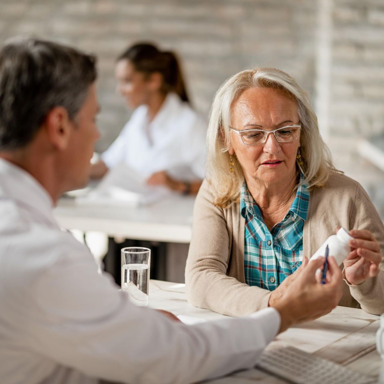 An older woman sits at a table talking to a doctor who is showing her a bottle of vitamins