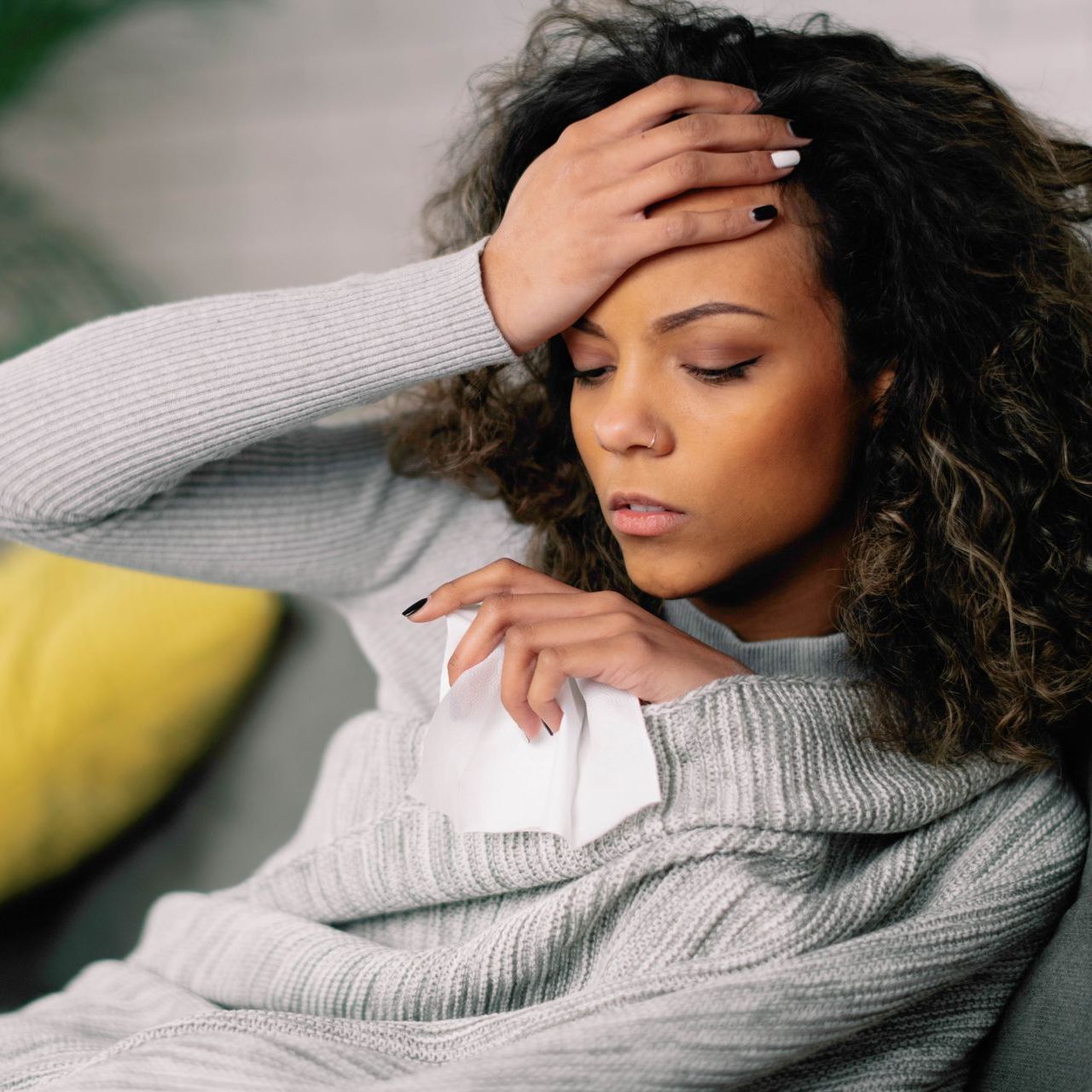 A woman sitting on a couch wrapped in a blanket holding a tissue and her forehead