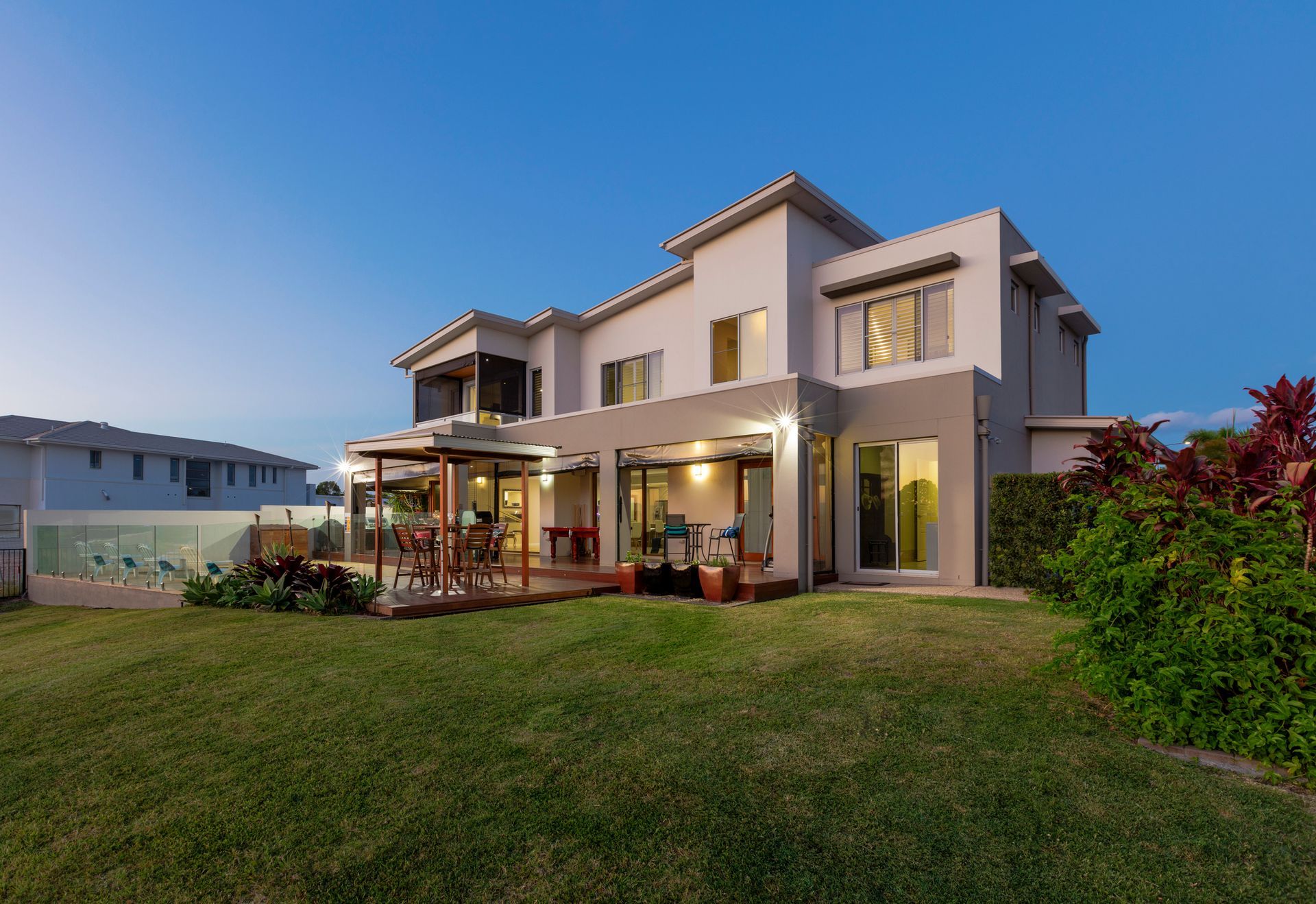 A modern two-story house at dusk featuring large windows, an outdoor patio with seating, a covered dining area