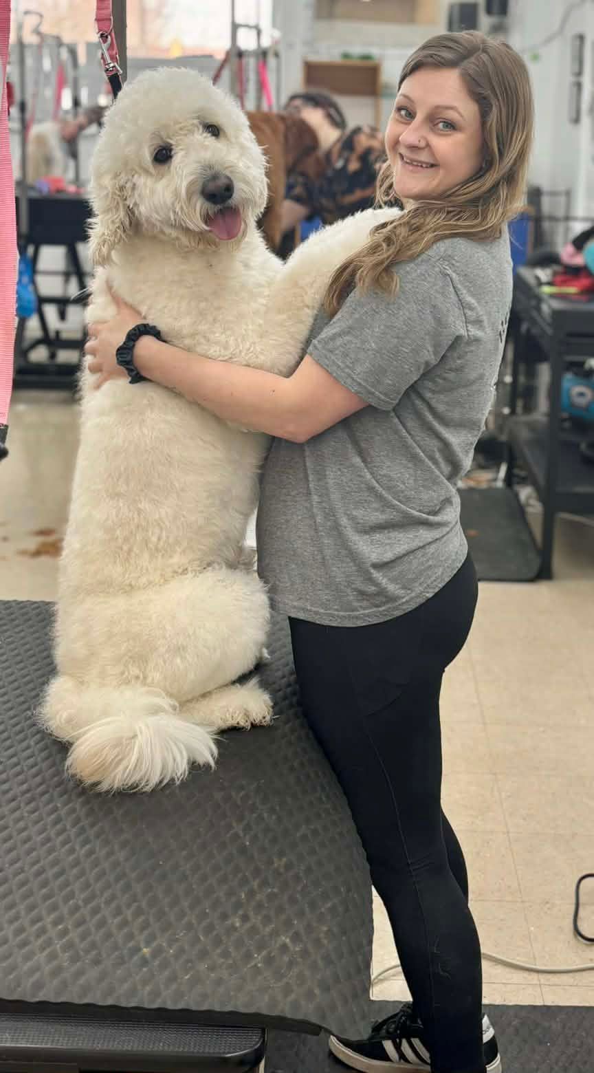 Woman hugging a large white dog on a grooming table. Dog is standing tall, tongue out.