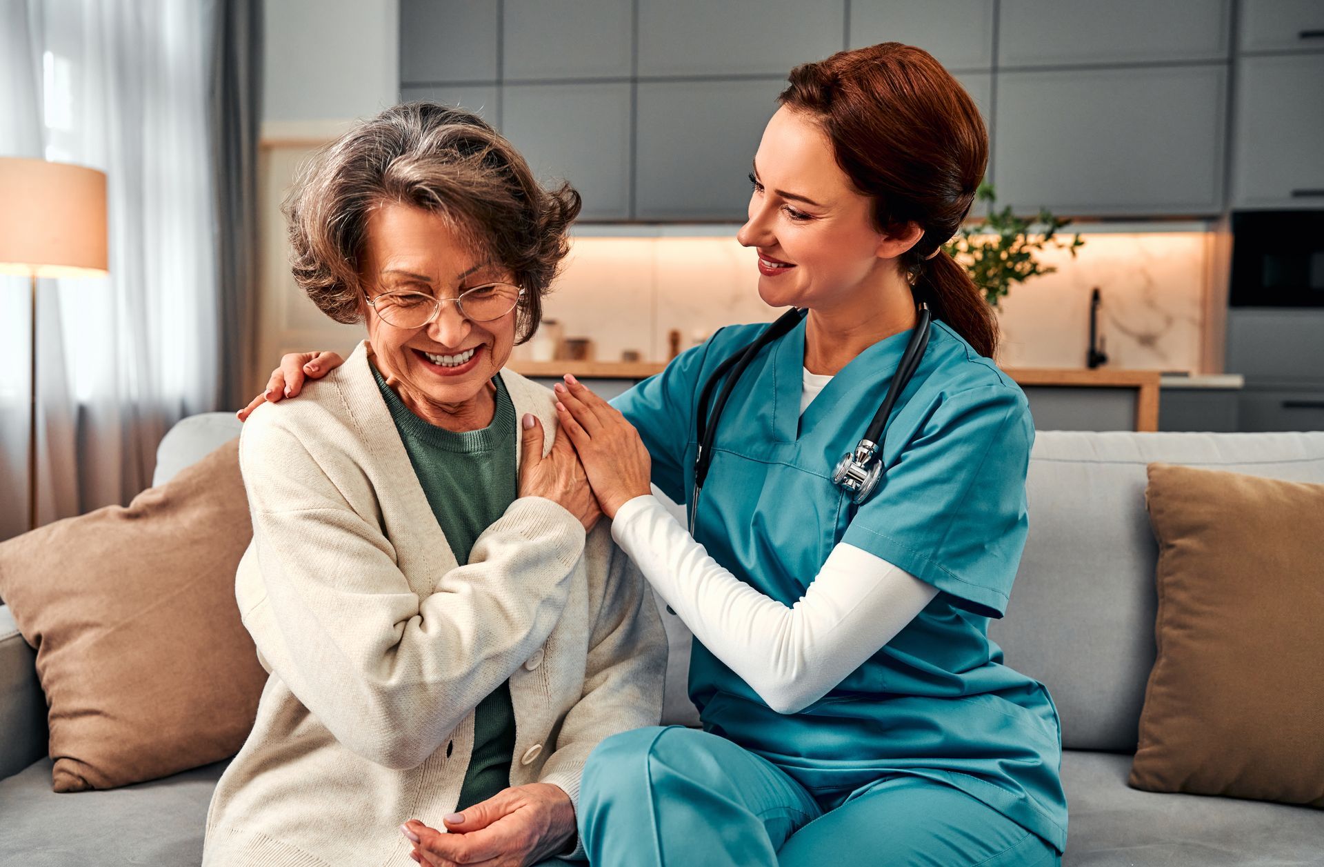 Nurse comforts older woman, both smiling, sitting on a couch. Interior setting.