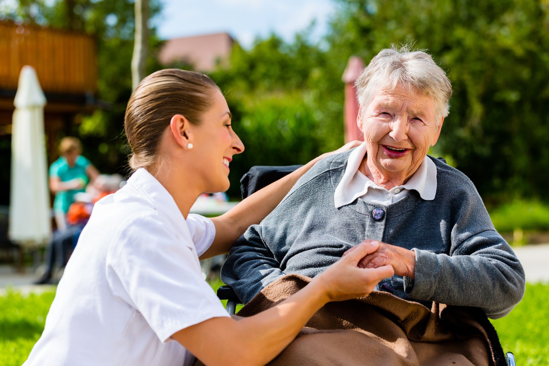 Caregiver in white uniform smiling and holding hands with smiling elderly woman in wheelchair outdoors.