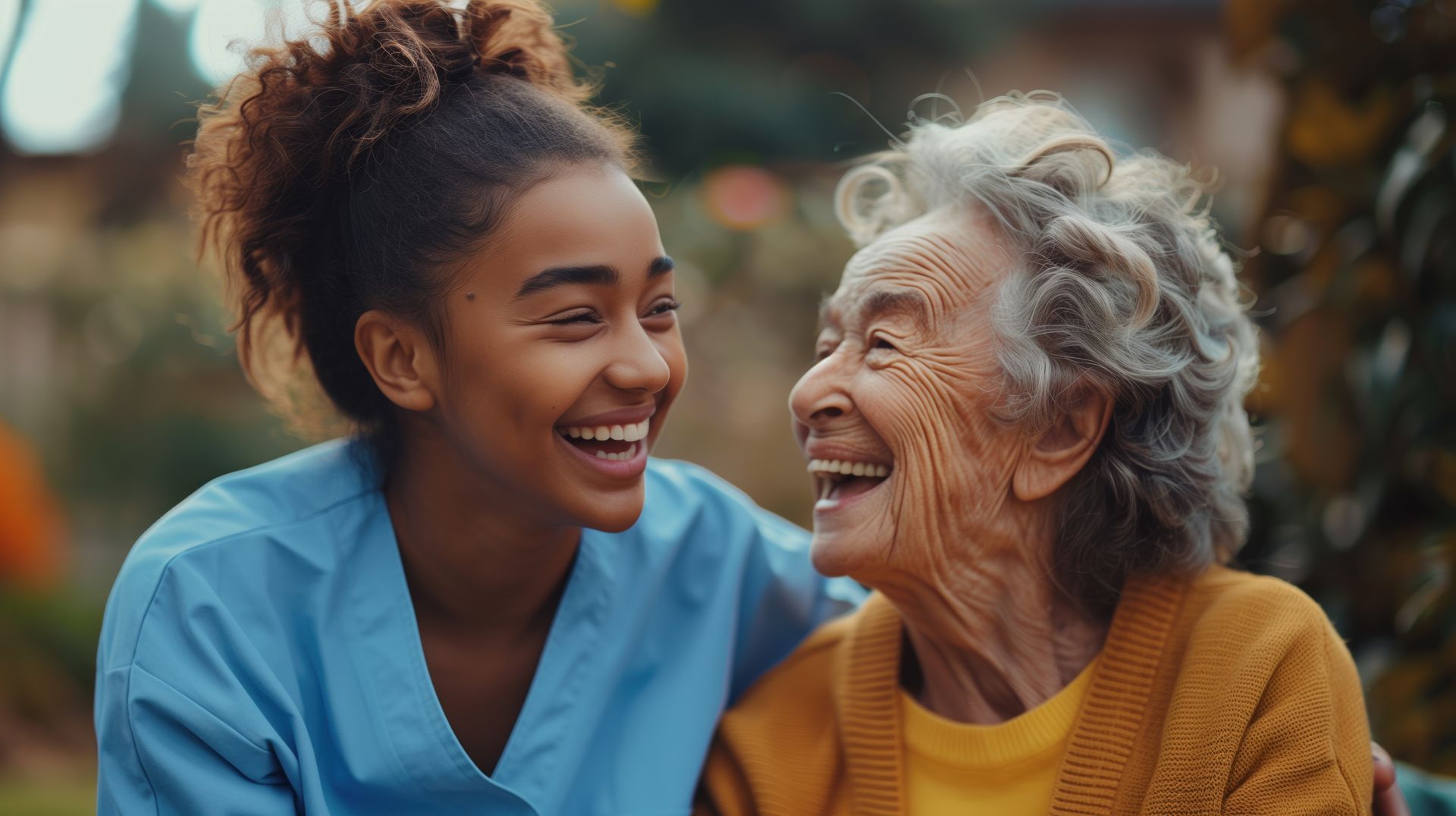 Young woman in blue scrubs smiles at elderly woman outdoors. Both laugh.