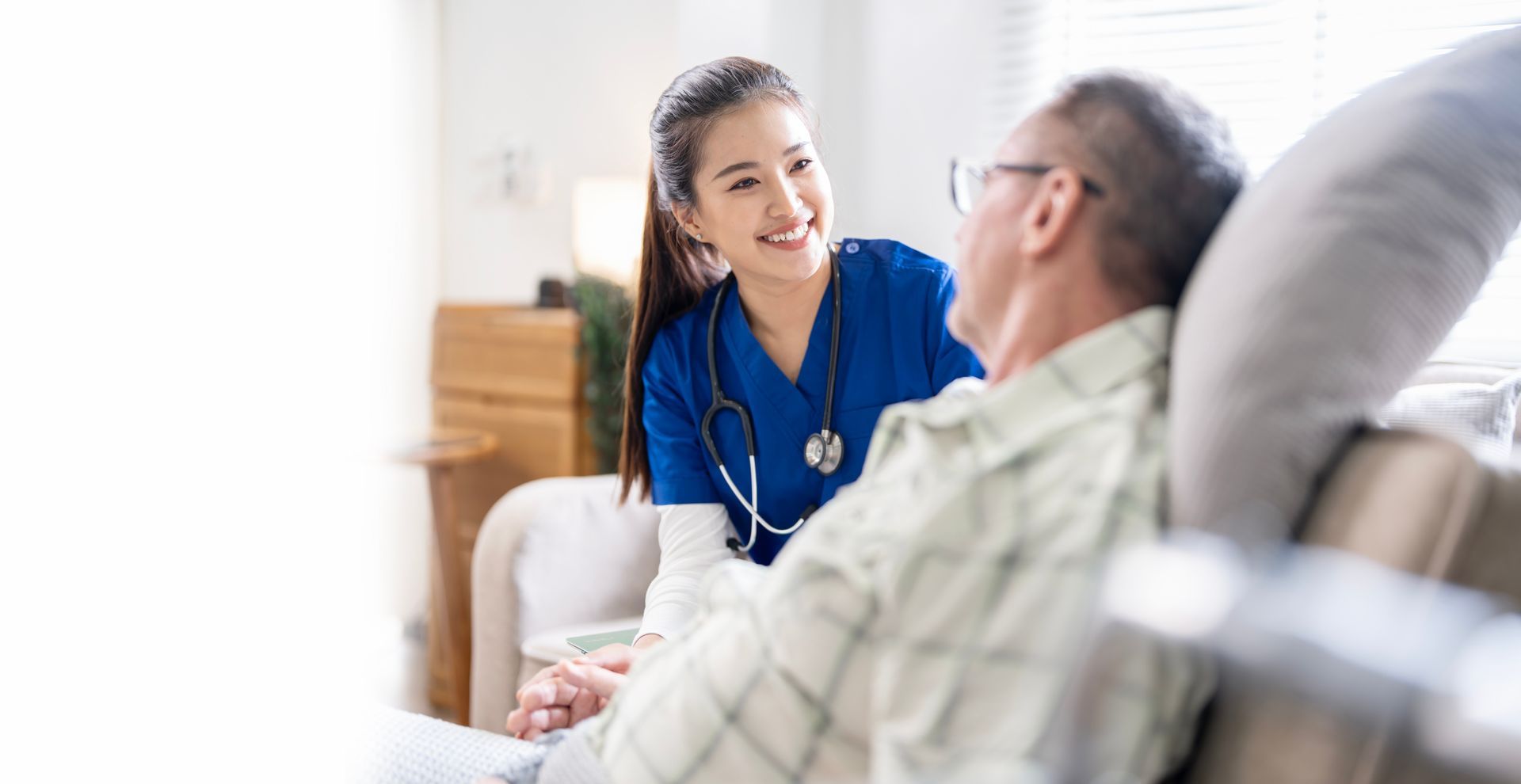 Nurse in blue scrubs smiles at a man in a plaid shirt seated on a sofa.