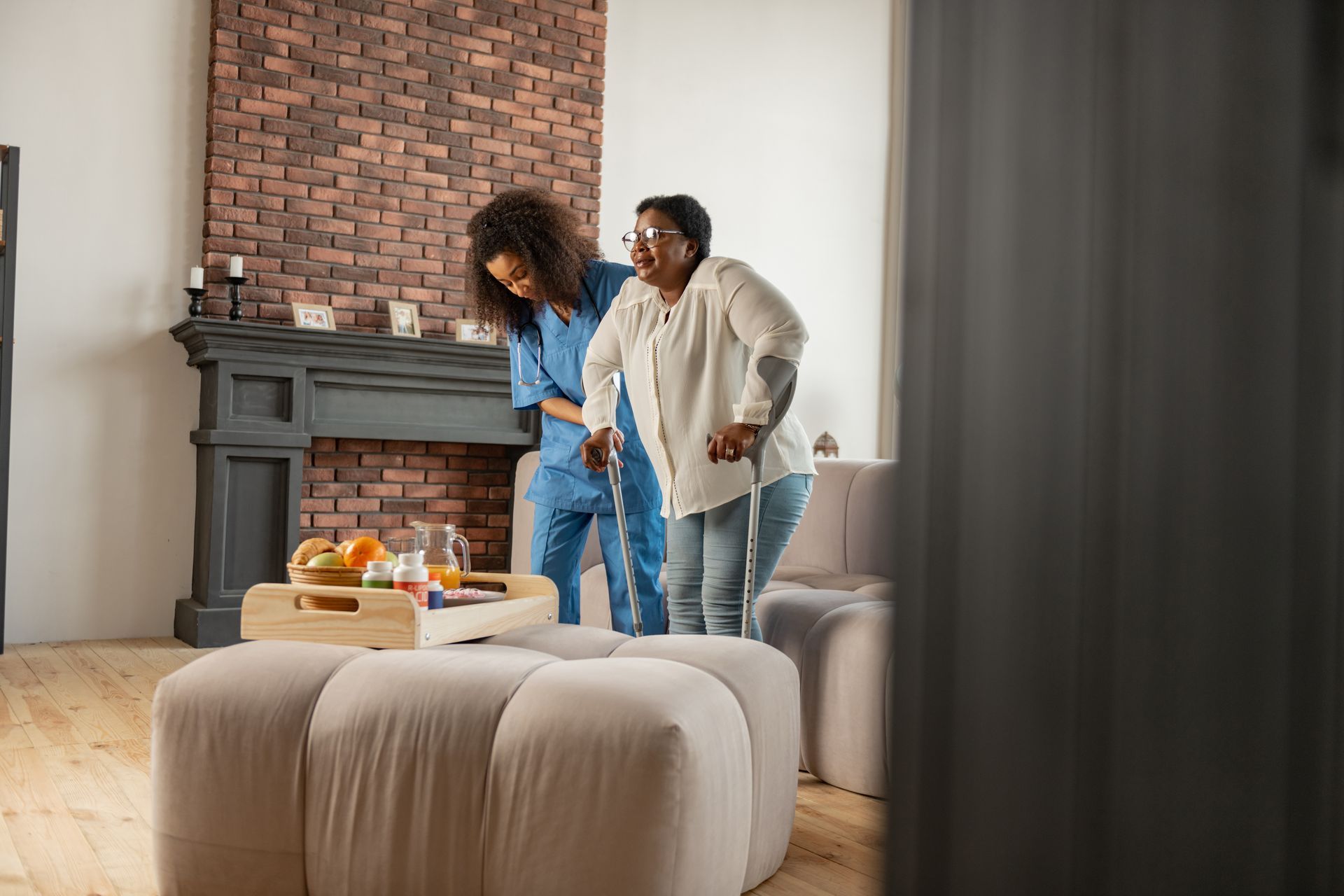 Caregiver assisting a person with mobility issues in a living room; a tray with medicine sits on the ottoman.