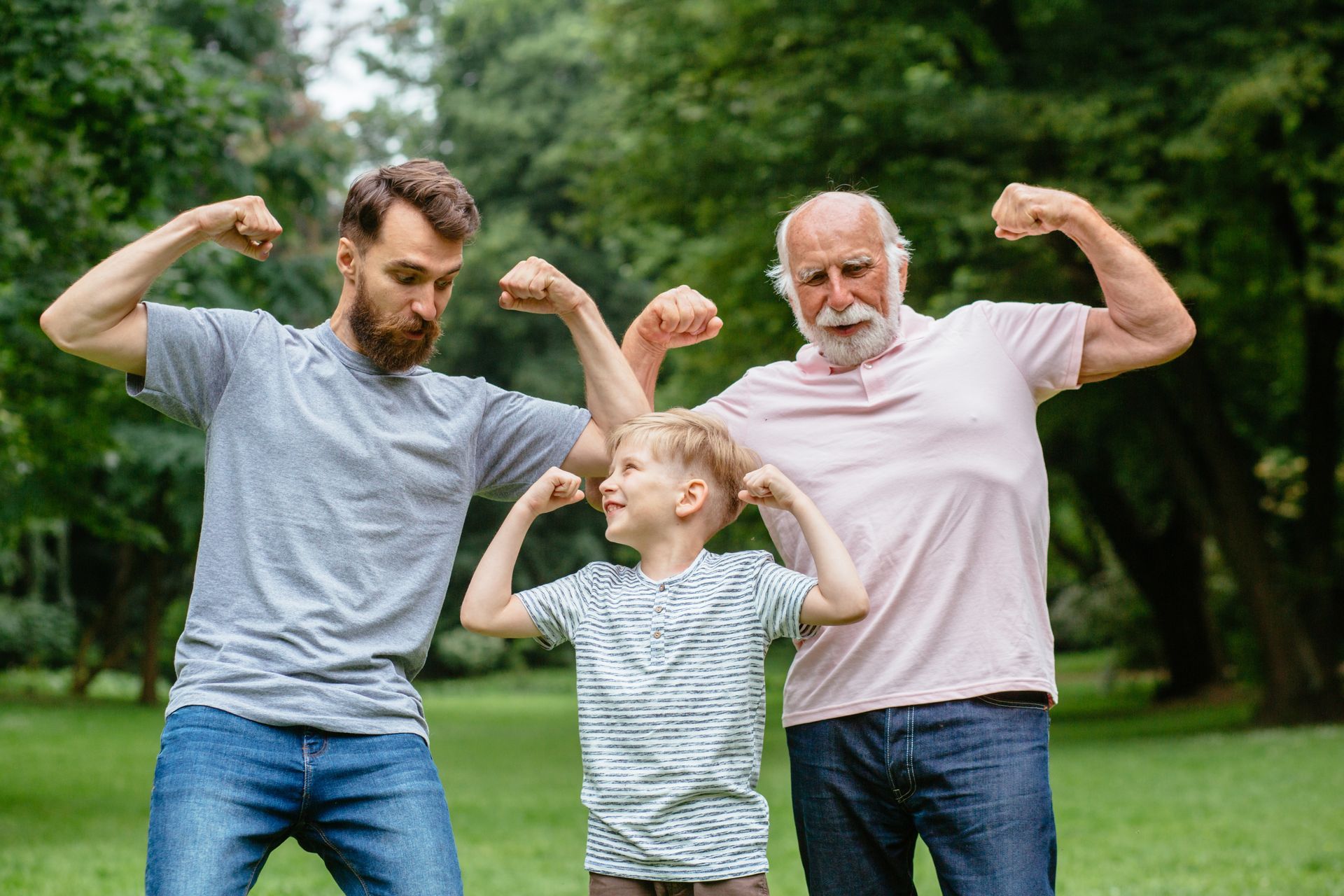 Three generations flexing biceps in a park: father, son, and grandfather.