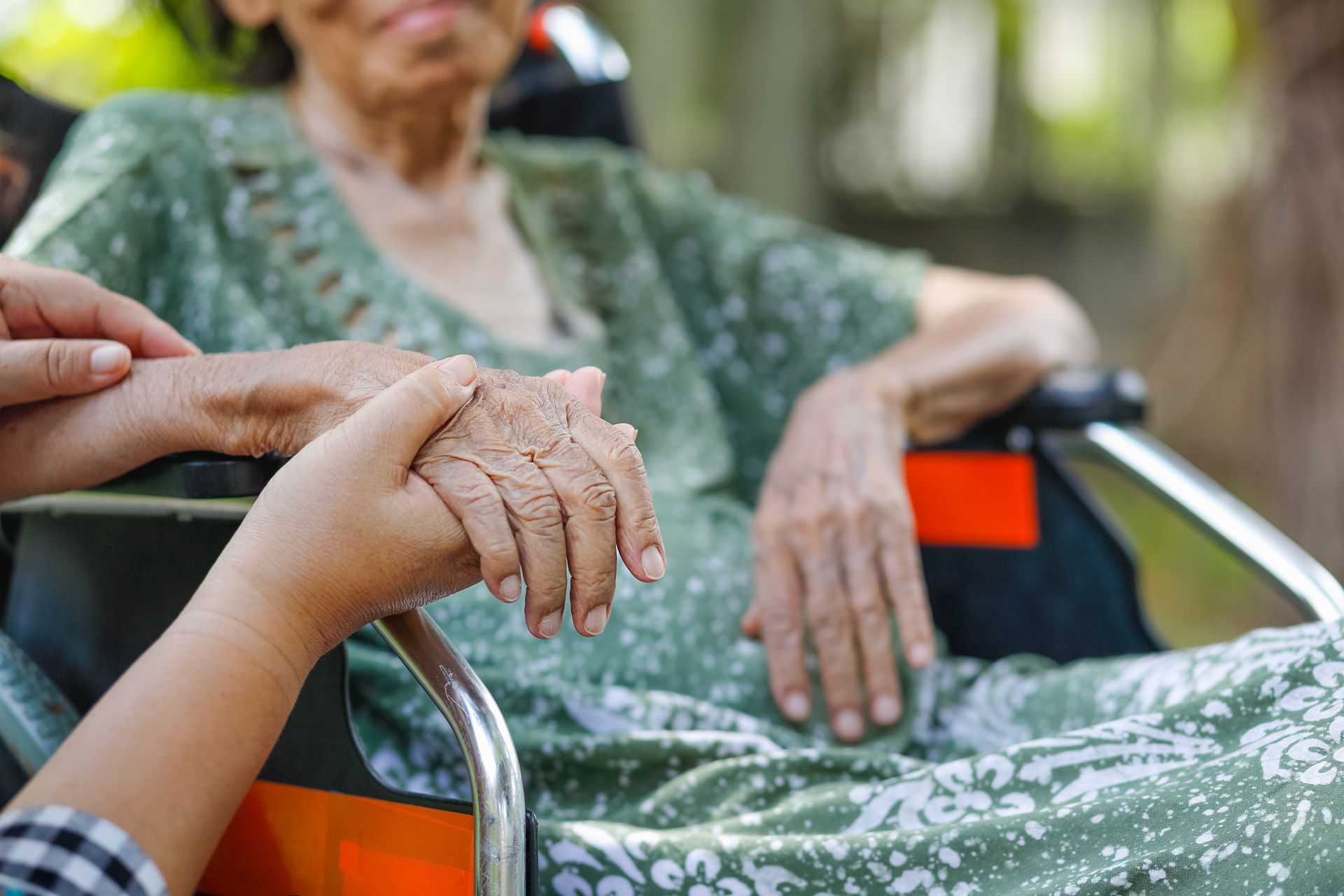 Person holding hands with a person in a wheelchair. Outdoors, soft green clothing.