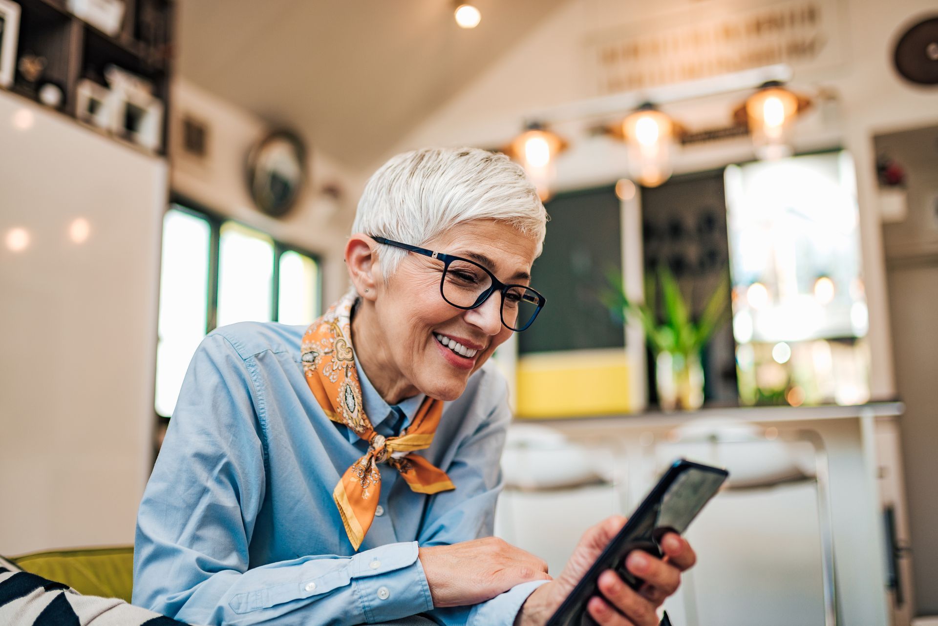 Woman with glasses smiles while using a tablet. She wears a blue shirt and orange scarf; interior setting.