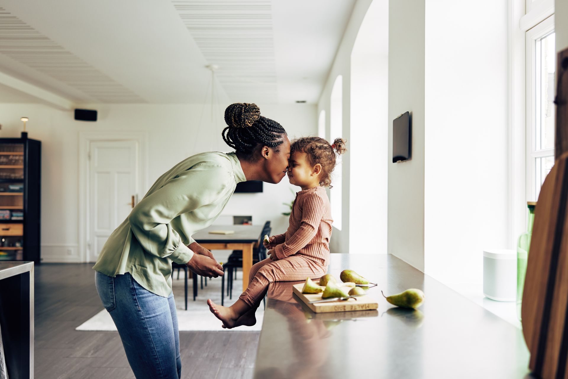 Woman kissing child on forehead, sitting on countertop in sunny kitchen.