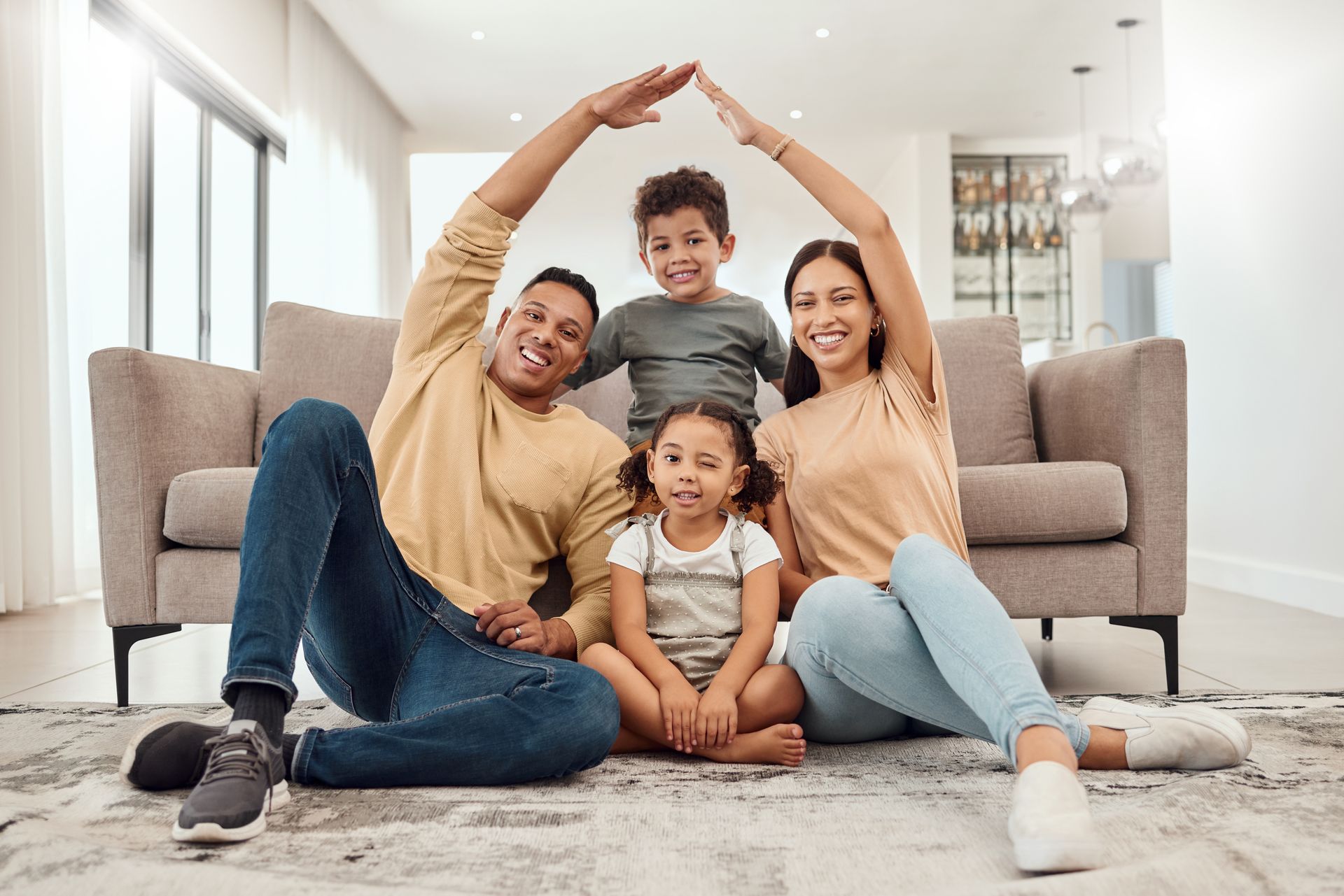 Family of four forms roof with hands, smiling near sofa.