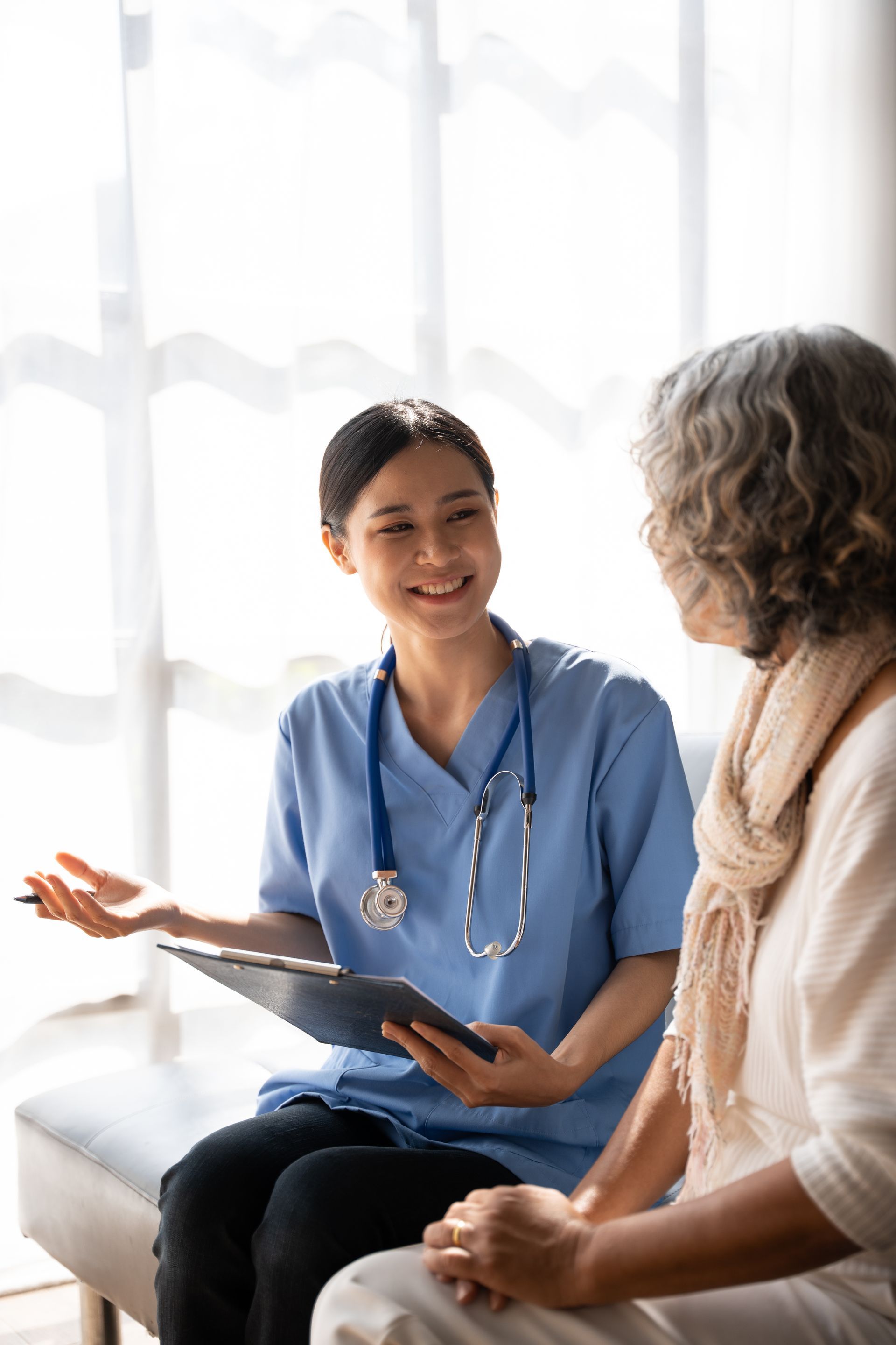 Nurse in blue scrubs with stethoscope, smiling, talking with a patient, holding a clipboard.