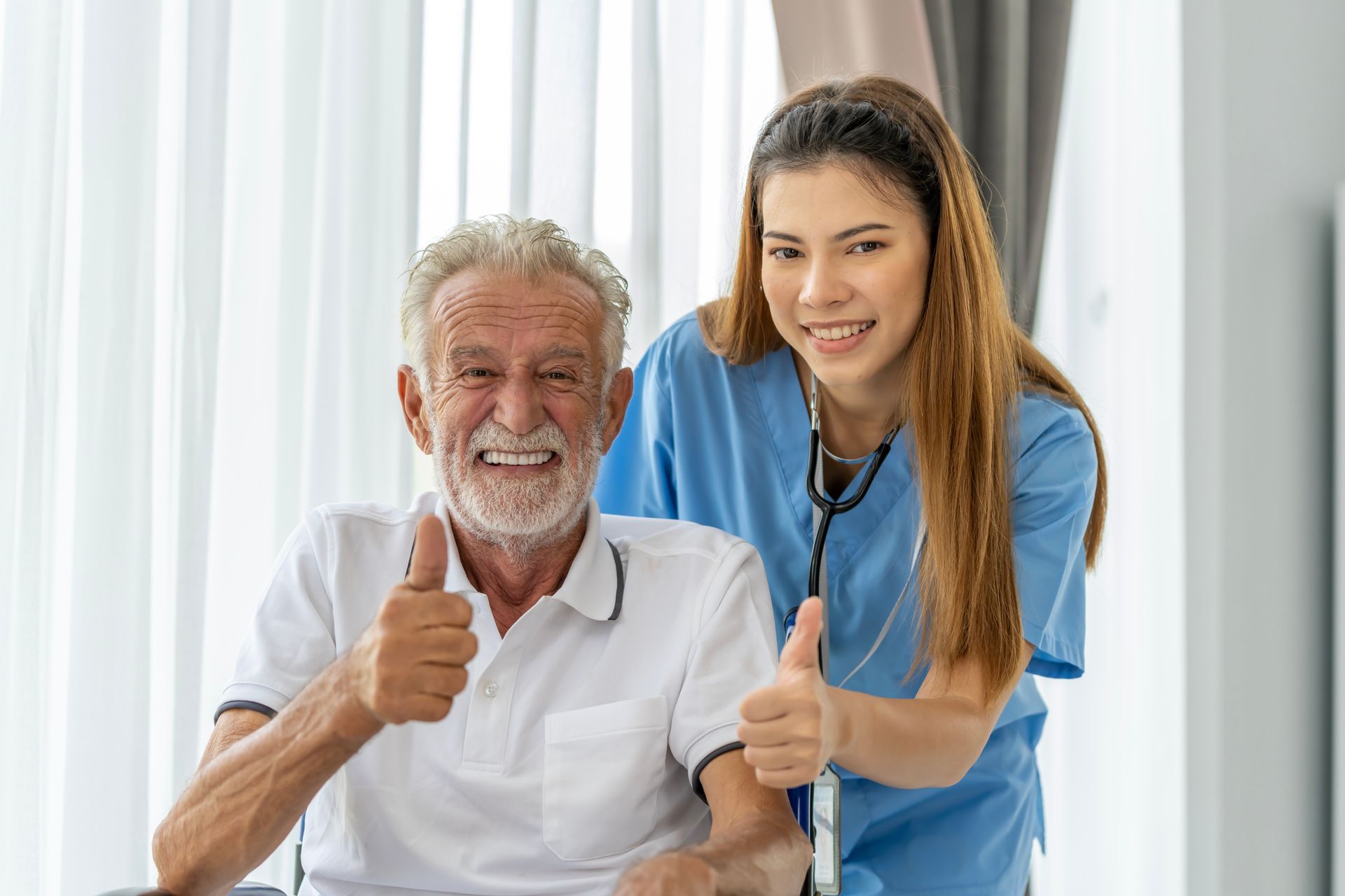 Smiling senior and caregiver giving thumbs up in a room with window.