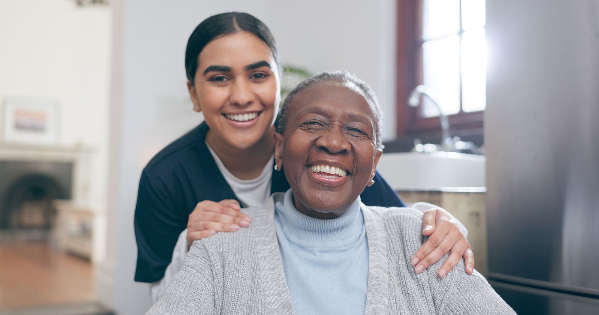 Woman in scrubs with hands on smiling elderly woman's shoulders, indoors.