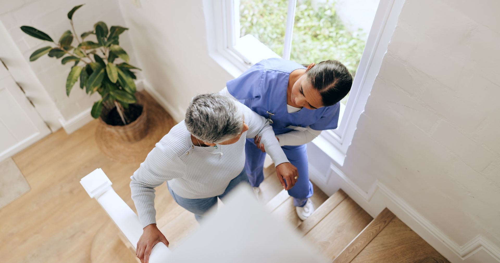 Caregiver assisting a person up stairs, holding their arm. They are near a window.