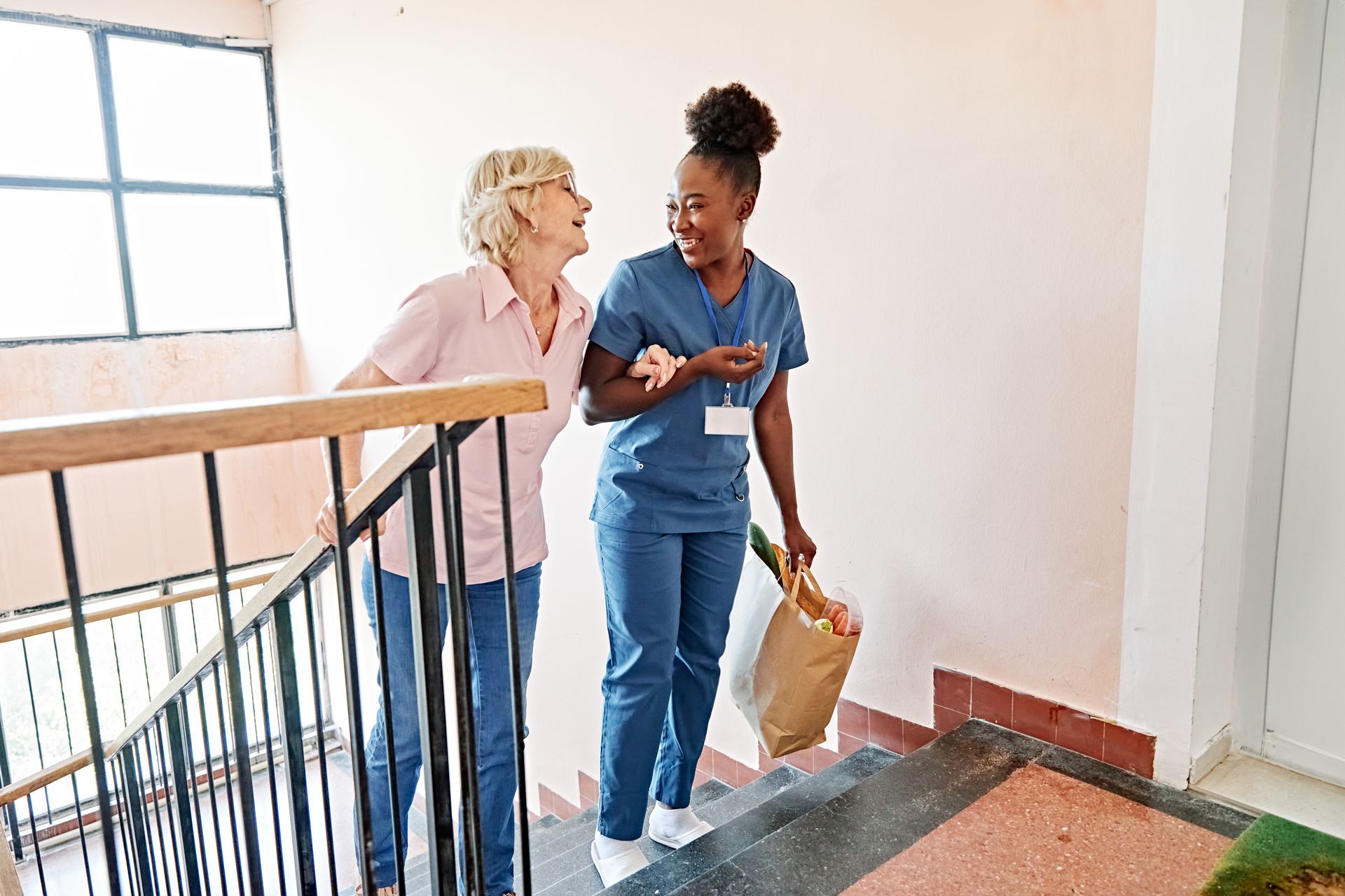 Caregiver helping someone down stairs, holding groceries; both are smiling.