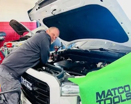 A mechanic in a gray hoodie leans over a white GMC truck's open hood, working on the engine. The Matco Tools logo is visible.