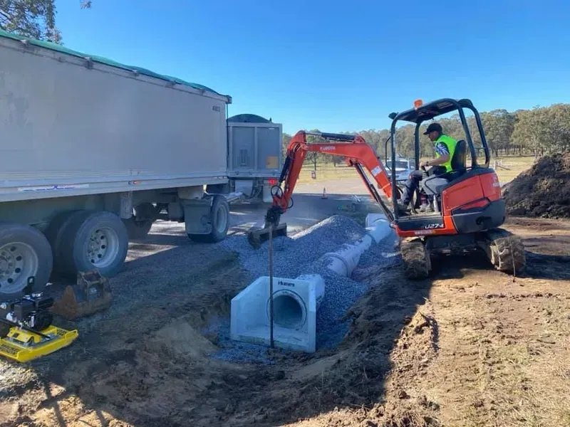 A Man Is Driving an Excavator on A Dirt Road Next to A Truck — Medowie Plumbing & Gas In Medowie, NSW