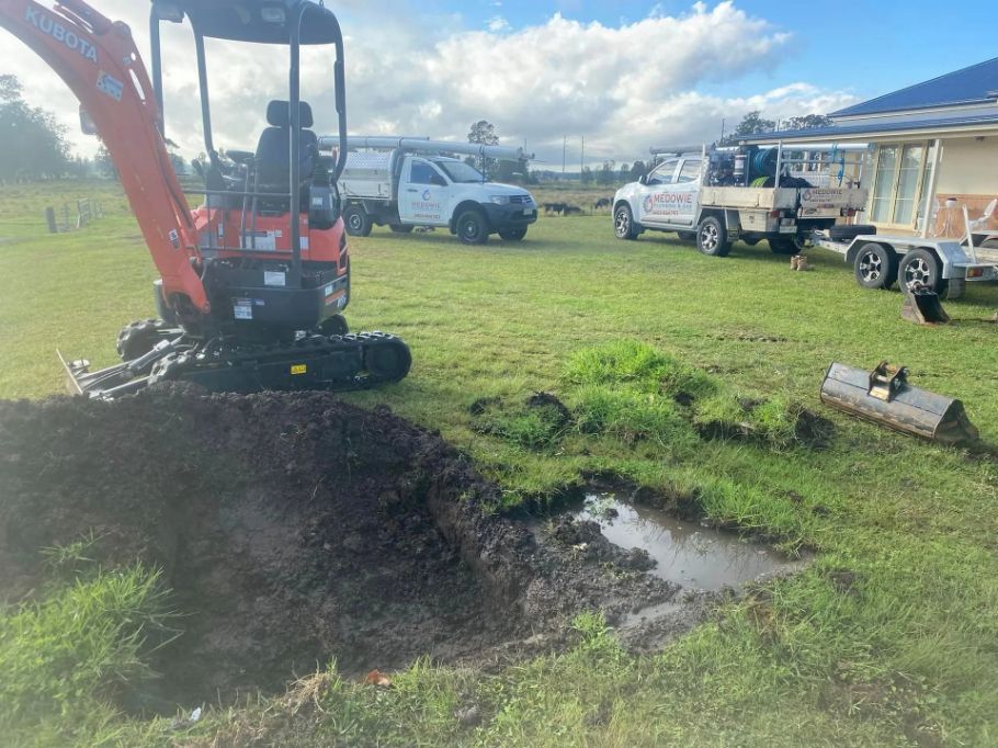 A Small Excavator Is Digging a Hole in The Grass in A Field — Medowie Plumbing & Gas In Medowie, NSW