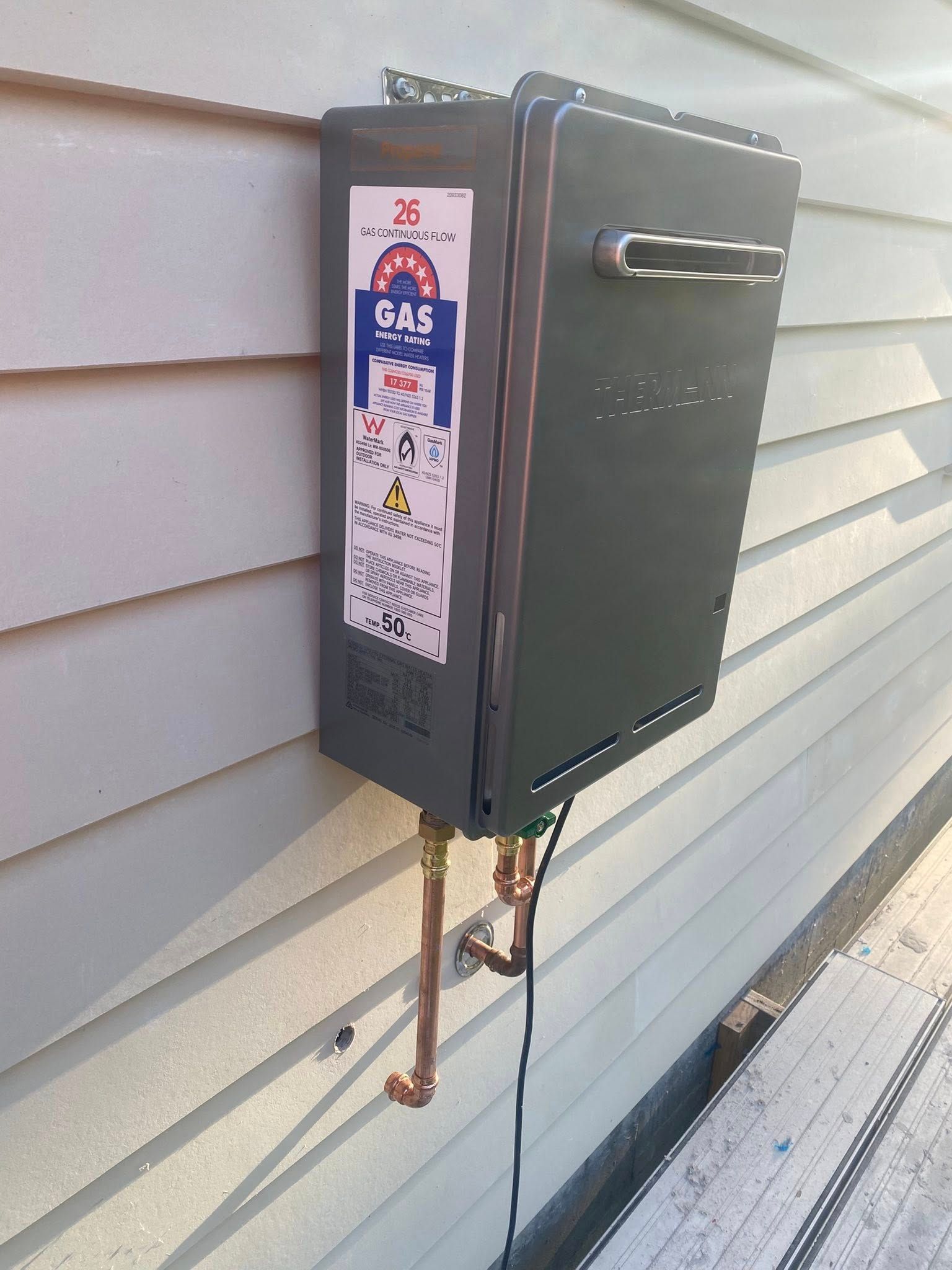 Dark Gray Tankless Water Heater Mounted on A Light Gray House Siding — Medowie Plumbing & Gas in in Newcastle