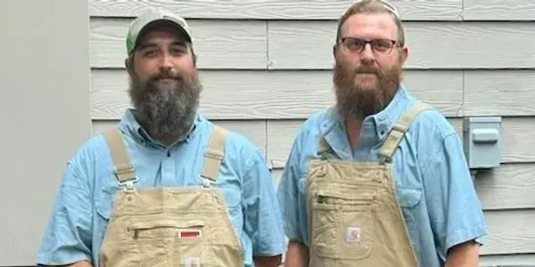 Two men with beards are standing next to each other wearing overalls.