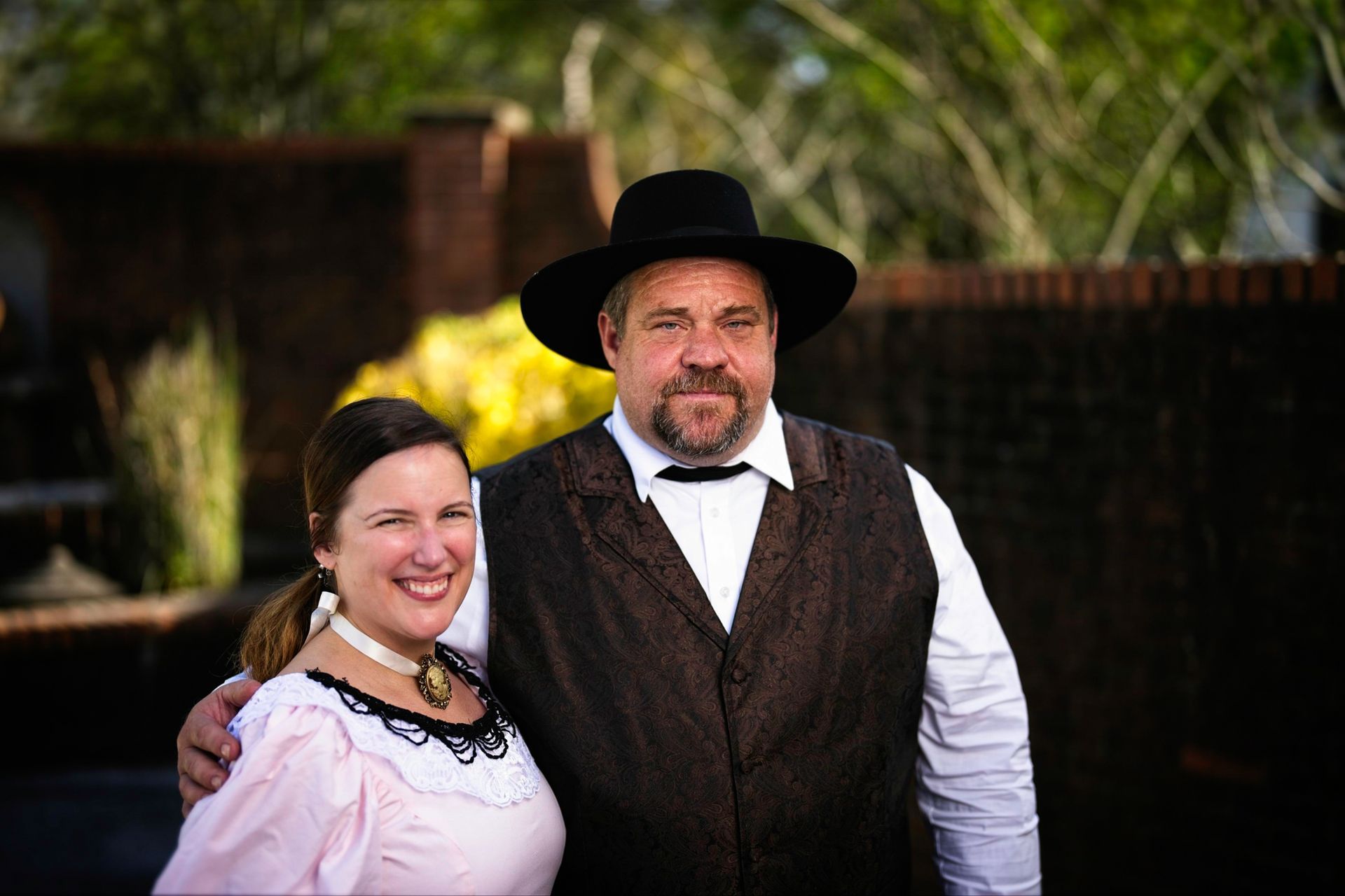 A man in a hat and vest is standing next to a woman in a pink dress.